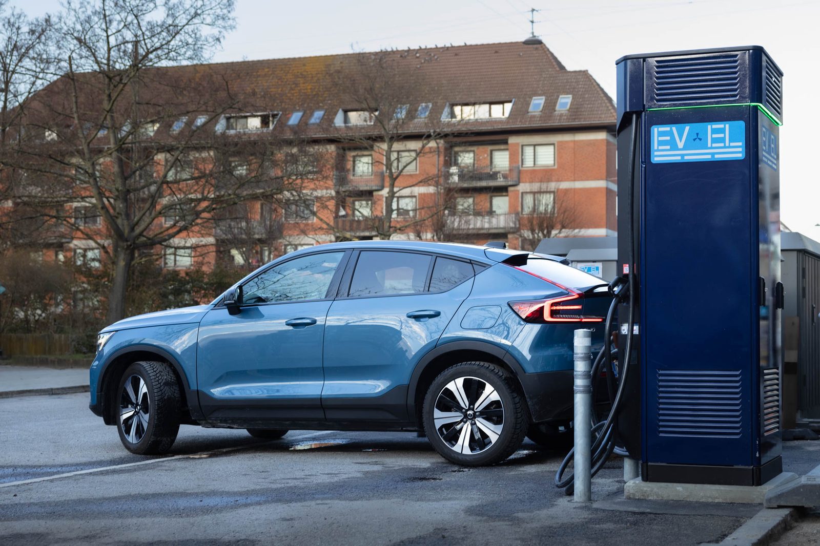 Blue electric car plugged into an EV-EL fast charger in front of a red brick building